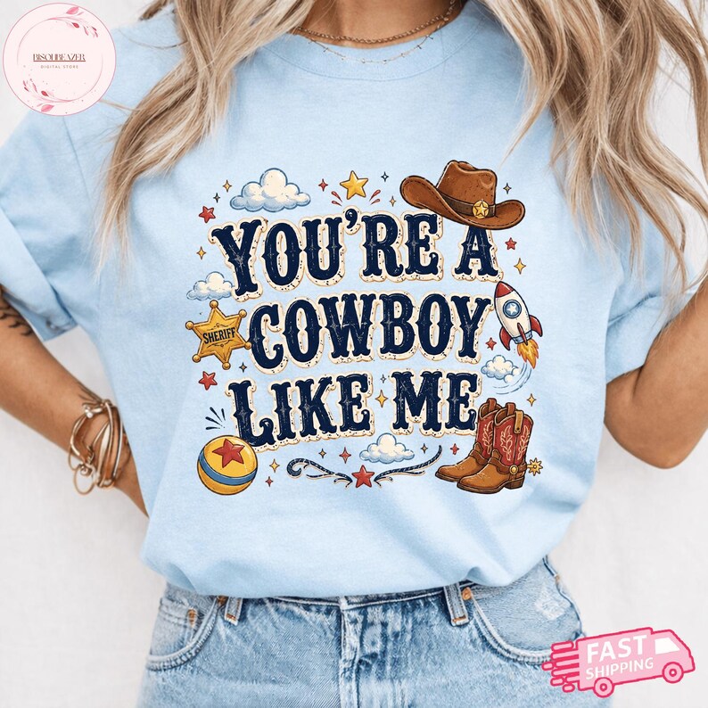 May include: A light blue t-shirt featuring the text "YOU'RE A COWBOY LIKE ME" in dark blue letters. The graphic includes a brown cowboy hat, a sheriff star badge, a rocket ship, a pair of brown cowboy boots, a yellow ball with a red star, and clouds.