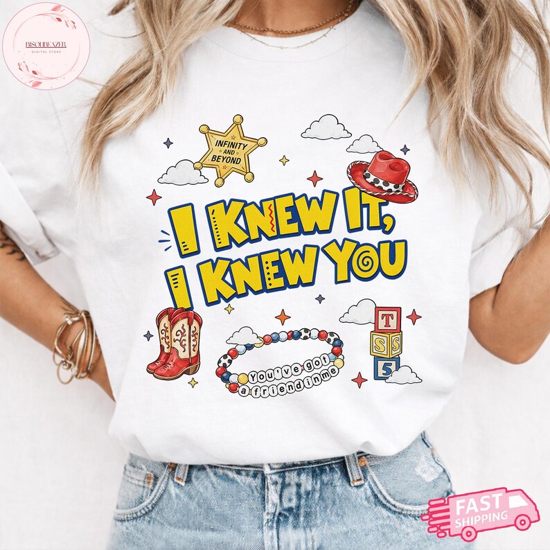 May include: A white t-shirt featuring a graphic print with the text I KNEW IT, I KNEW YOU in yellow and blue. The design includes a yellow sheriff star, a red cowboy hat, red cowboy boots, a beaded bracelet with the text YOU'VE GOT A FRIEND IN ME, and stacked alphabet blocks.