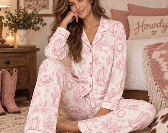 May include: A two-piece pajama set featuring a long-sleeved button-down shirt and matching pants in a white fabric with a pink western-themed print. The pattern includes illustrations of cowboy hats, boots, lassos, and cacti with pink piping.