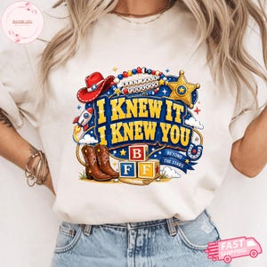 May include: A cream-colored t-shirt featuring a graphic print with the text I KNEW IT I KNEW YOU, BEST PALS FOREVER, and BEYOND THE STARS. The design includes a red cowboy hat, a gold sheriff star, a rocket, brown cowboy boots, and alphabet blocks.