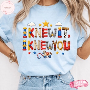 Puede incluir: Una camiseta azul claro con un dise&ntilde;o gr&aacute;fico que incluye el texto "I KNEW IT, I KNEW YOU" en letras coloridas inspiradas en juguetes. El dise&ntilde;o presenta peque&ntilde;as estrellas, nubes y un caballo de juguete para arrastrar, todo en un estilo l&uacute;dico.