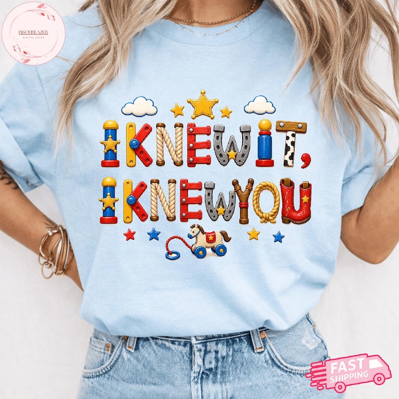 Puede incluir: Una camiseta azul claro con un dise&ntilde;o gr&aacute;fico que incluye el texto "I KNEW IT, I KNEW YOU" en letras coloridas inspiradas en juguetes. El dise&ntilde;o presenta peque&ntilde;as estrellas, nubes y un caballo de juguete para arrastrar, todo en un estilo l&uacute;dico.