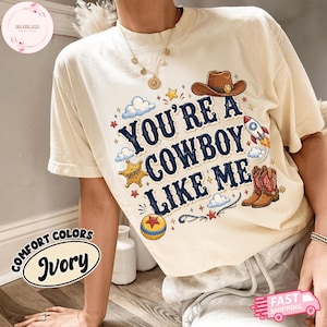 May include: An ivory t-shirt featuring the text "YOU'RE A COWBOY LIKE ME" in dark blue letters. The graphic includes a brown cowboy hat, a yellow sheriff star badge, a colourful rocket, a pair of brown cowboy boots, and a yellow and red ball.