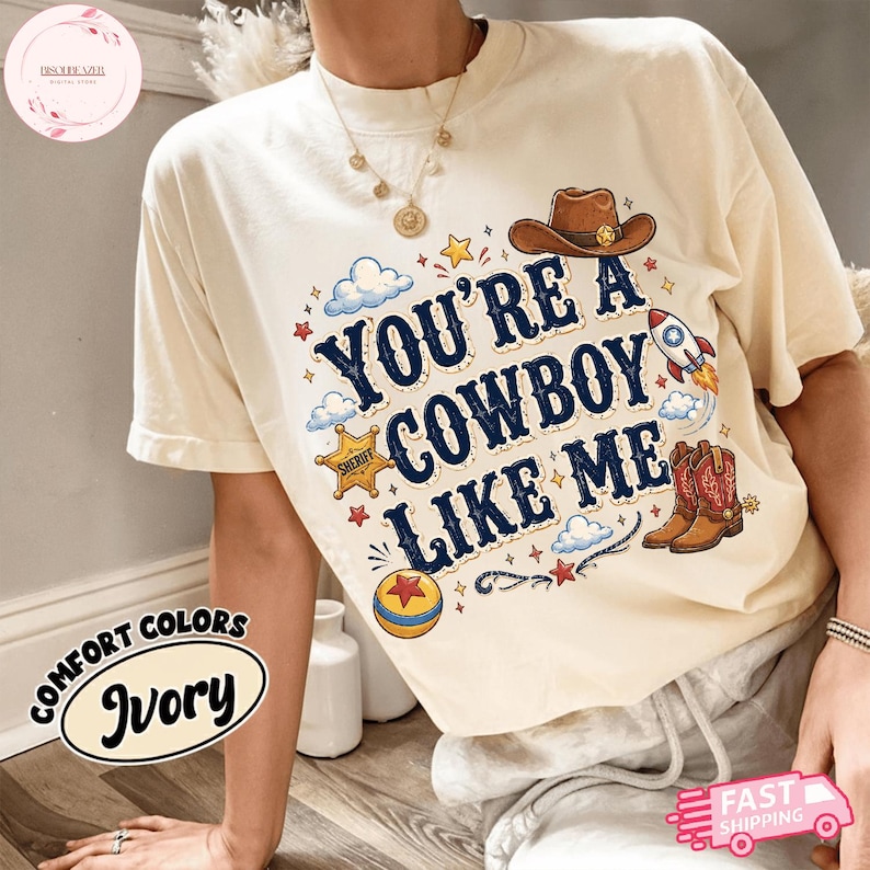 May include: An ivory t-shirt featuring the text "YOU'RE A COWBOY LIKE ME" in dark blue letters. The graphic includes a brown cowboy hat, a yellow sheriff star badge, a colourful rocket, a pair of brown cowboy boots, and a yellow and red ball.