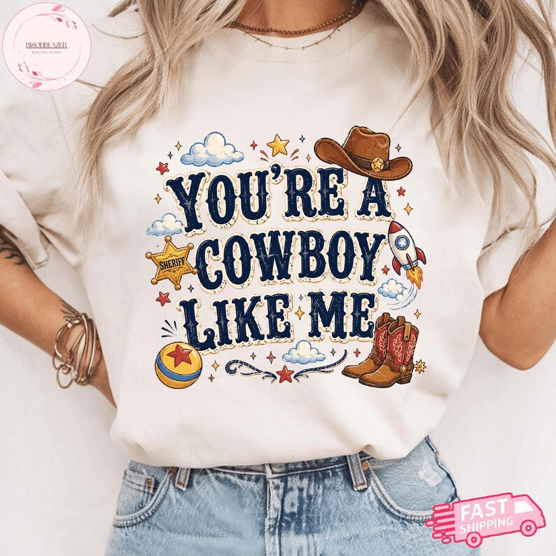 May include: A cream-coloured t-shirt featuring the dark blue text "YOU'RE A COWBOY LIKE ME" surrounded by colourful illustrations including a brown cowboy hat, a sheriff star badge, a rocket ship, a red and yellow ball, and a pair of cowboy boots.