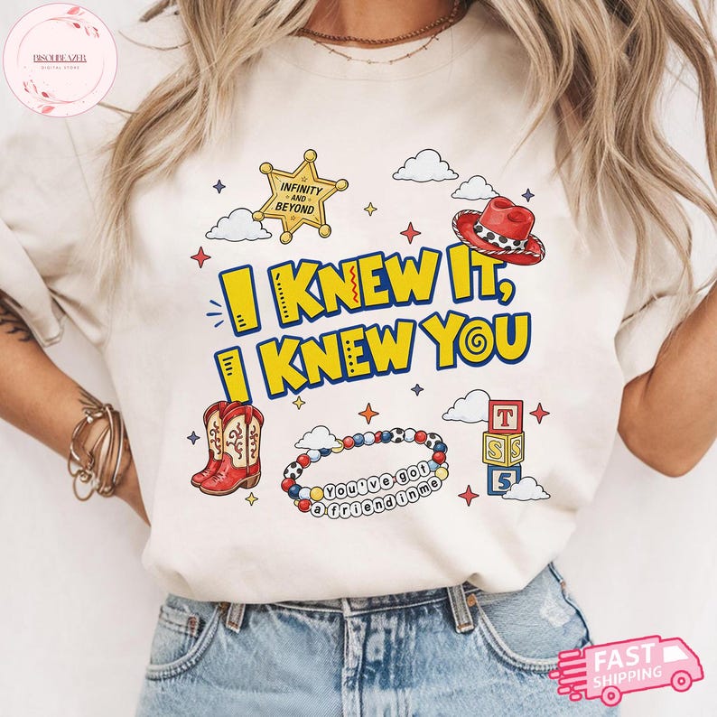 May include: A cream-colored t-shirt featuring a graphic print with the text I KNEW IT, I KNEW YOU. The design includes a yellow sheriff star, a red cowboy hat, red cowboy boots, a beaded bracelet with the text You've got a friend in me, and blocks.