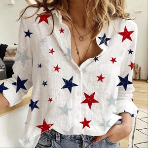 May include: A white long-sleeve button-down shirt featuring a scattered pattern of red, navy blue, and light gray stars. The shirt has a collared neckline, a front button placket, and long sleeves with cuffs, showcasing a textured fabric finish.