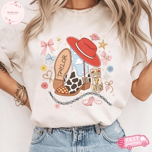 May include: A cream-colored t-shirt featuring a graphic print of a cow-print cowboy boot, a red cowboy hat, building blocks with the letters T, S, and the number 13, pink bows, flowers, and a beaded necklace with the text THE NEW YOU.