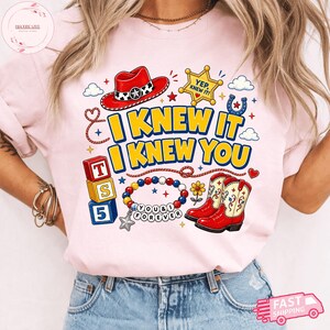 Pode incluir: Uma t-shirt rosa claro com uma estampa gr&aacute;fica com o texto I KNEW IT I KNEW YOU. O design inclui um chap&eacute;u de cowboy vermelho, uma estrela de xerife amarela, blocos coloridos, uma pulseira de contas com o texto YOU & I FOREVER e botas vermelhas.