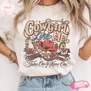 Op de afbeelding: Een cr&egrave;mekleurig t-shirt met een grafische print met de tekst Cowgirl Like Me en Takes One to Know One. Het ontwerp bevat een roze cowboyhoed, een hoefijzer, een hobbelpaard, een sheriffster, rozen, muzieknoten en een lasso.