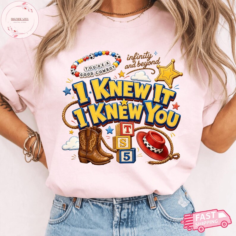May include: A light pink t-shirt featuring a graphic print with the text "I KNEW IT I KNEW YOU" and "infinity and beyond". The design includes a beaded bracelet, a yellow sheriff star, brown cowboy boots, alphabet blocks, and a red hat.