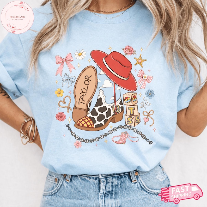 May include: A light blue t-shirt featuring a graphic print of a red cowboy hat, a cow-print cowboy boot with the name TAYLOR and the number 13, stacked blocks with the letters T, S, and a heart, surrounded by bows, flowers, and stars.
