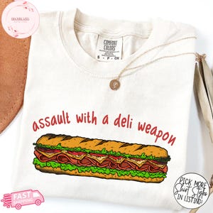 Könnte beinhalten: Cremefarbenes T-Shirt mit dem roten Schriftzug "assault with a deli weapon" und einer Abbildung eines Sandwiches. Das Shirt hat ein Comfort Colors-Etikett und eine goldene Halskette.