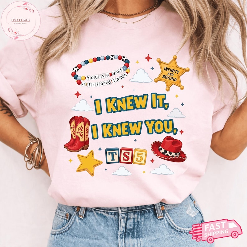 May include: A light pink t-shirt featuring a graphic print with the text I KNEW IT, I KNEW YOU, TS5, INFINITY AND BEYOND, and YOU'VE GOT A FRIEND IN ME. The design includes a beaded bracelet, a sheriff star badge, red cowboy boots, a red hat, and stars.