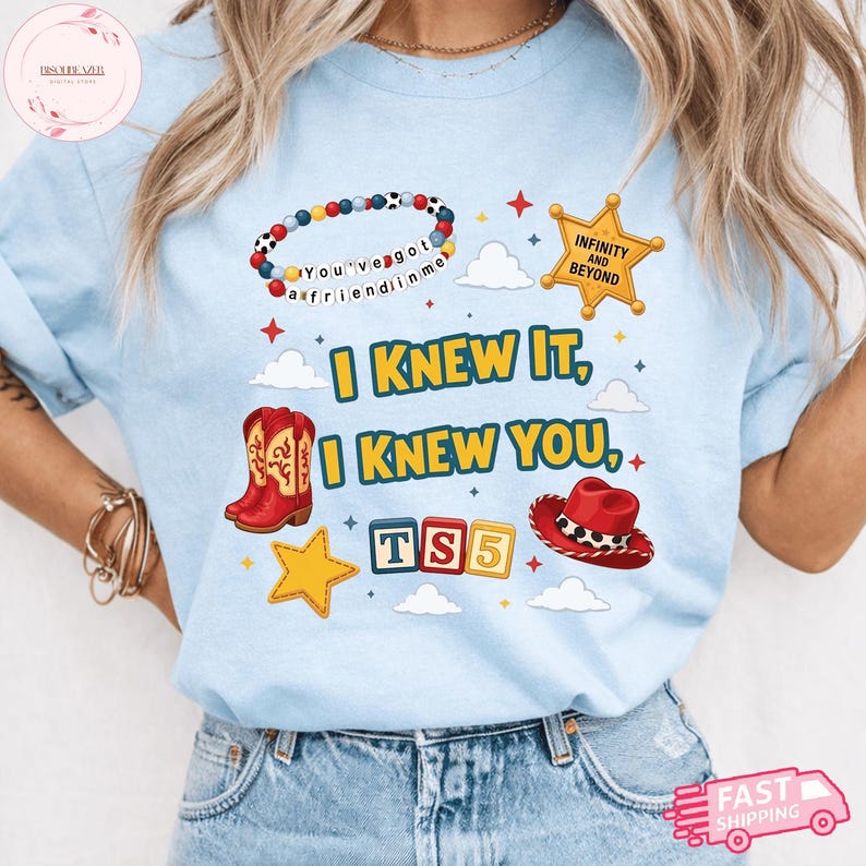 May include: A light blue t-shirt featuring a graphic print with the text I KNEW IT, I KNEW YOU, TS5, INFINITY AND BEYOND, and YOU'VE GOT A FRIEND IN ME. The design includes a beaded bracelet, a sheriff star, red cowboy boots, a red hat, and stars.