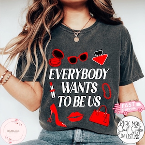 May include: Dark gray t-shirt with the text "EVERYBODY WANTS TO BE US" in white. The shirt features red illustrations of makeup, sunglasses, perfume, lipstick, a necklace, a high heel, a pair of lips, and a handbag.