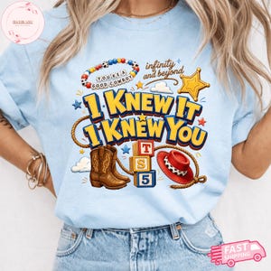 May include: A light blue t-shirt featuring a graphic print with brown cowboy boots, a red cowboy hat, a yellow sheriff star, alphabet blocks, and a beaded bracelet. The text reads "YOU'RE A GOOD COWBOY", "infinity and beyond", "I KNEW IT", and "I KNEW YOU".