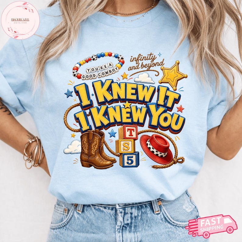 May include: A light blue t-shirt featuring a graphic print with brown cowboy boots, a red cowboy hat, a yellow sheriff star, alphabet blocks, and a beaded bracelet. The text reads "YOU'RE A GOOD COWBOY", "infinity and beyond", "I KNEW IT", and "I KNEW YOU".