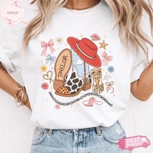 May include: A white t-shirt featuring a graphic print of a cow-print cowboy boot with the number 13, a red cowboy hat, alphabet blocks spelling TS, and the name Taylor. The design includes pink bows, flowers, stars, and a beaded string.
