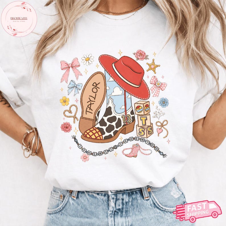 May include: A white t-shirt featuring a graphic print of a cow-print cowboy boot with the number 13, a red cowboy hat, alphabet blocks spelling TS, and the name Taylor. The design includes pink bows, flowers, stars, and a beaded string.
