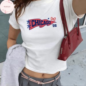 Puede incluir: Camiseta blanca con un diseño de bandera de Chicago en rojo y azul. La bandera muestra la palabra "CHICAGO" y el número "'26". La camiseta se combina con pantalones grises y un bolso rojo.