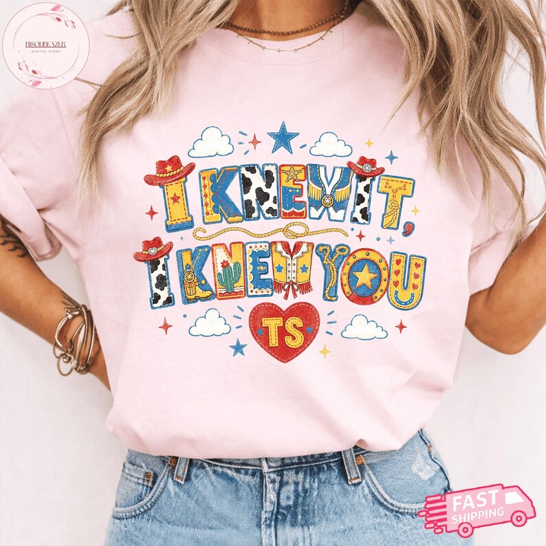 May include: A light pink t-shirt featuring a graphic print with the text I KNEW IT, I KNEW YOU and TS inside a red heart. The design includes cowboy hats, cow print patterns, stars, clouds, a cactus, and rope motifs in blue, yellow, and red.