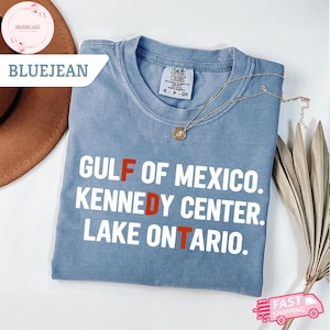 Op de afbeelding: Een jeansblauw t-shirt met korte mouwen en witte tekst die GULF OF MEXICO, KENNEDY CENTER, LAKE ONTARIO leest, waarbij de letters F, D en T in het rood zijn gemarkeerd. Het shirt heeft een ronde hals en een Comfort Colors merklabel.