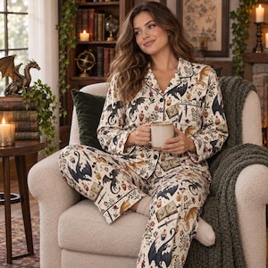 Puede incluir: Un conjunto de pijama de dos piezas con camisa de manga larga abotonada y pantalón a juego. La tela es de color crema con un patrón repetitivo de dragones negros y dorados, libros abiertos, dagas y frascos de pociones, con ribetes negros.