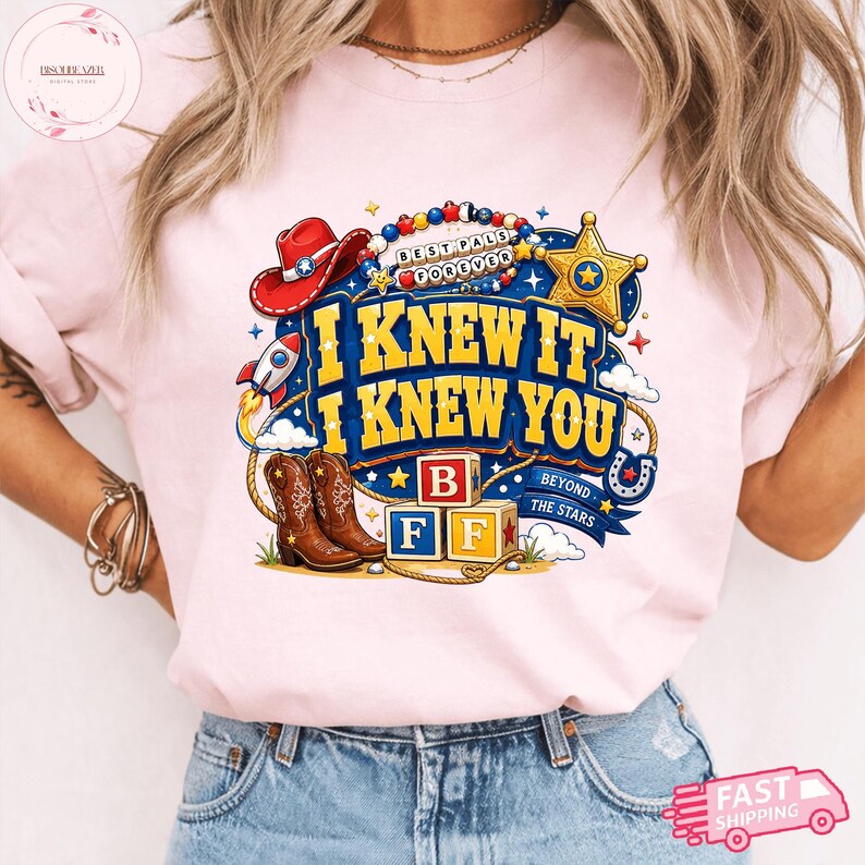 May include: A light pink t-shirt featuring a graphic print with the text I KNEW IT I KNEW YOU, BEST PALS FOREVER, and BEYOND THE STARS. The design includes a red cowboy hat, a gold sheriff badge, a rocket, brown cowboy boots, and alphabet blocks.