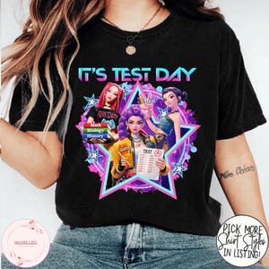 Op de afbeelding: Zwart T-shirt met de tekst "IT'S TEST DAY" in blauw en roze. De afbeelding toont drie anime-achtige figuren in een ster, die boeken, potloden en een testpapier vasthouden. Het shirt heeft ook de tekst "Math, Biology, History".