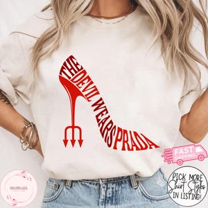 May include: Cream-colored t-shirt featuring a red high-heeled shoe design with the text "THE DEVIL WEARS PRADA". The shoe has a trident heel. Additional text includes "FAST SHIPPING" and "PICK MORE SHIRT STYLES IN LISTING!"