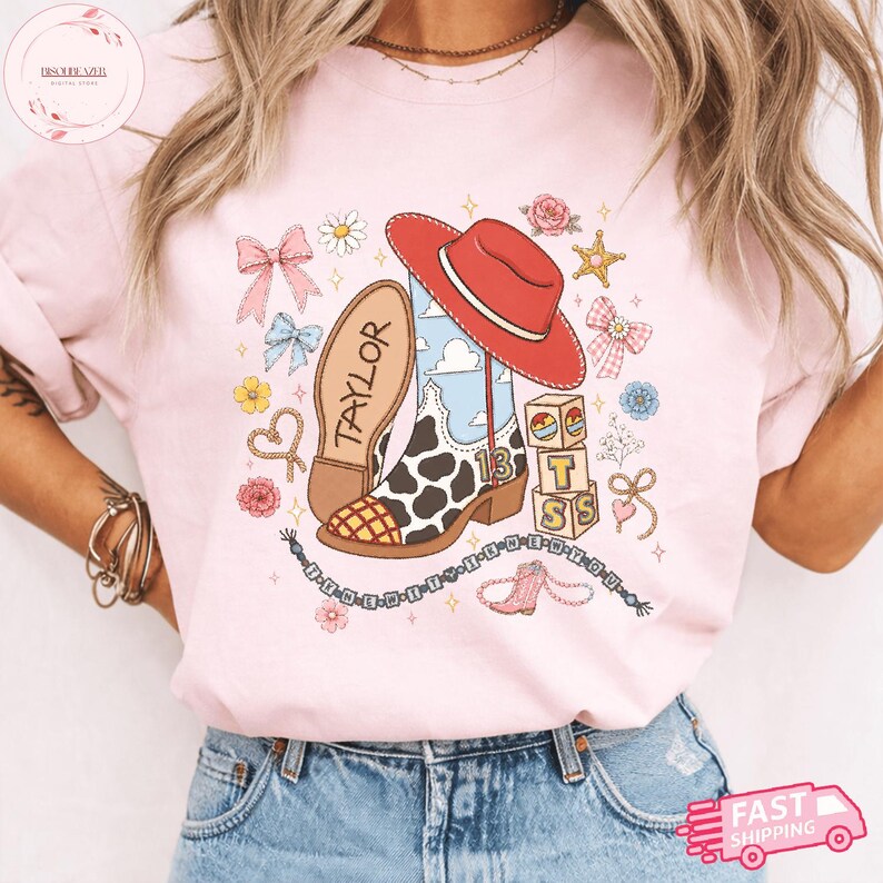 May include: A light pink t-shirt featuring a graphic print of a red cowboy hat, a cow-print boot with the number 13, and blocks spelling T S. The design includes bows, flowers, a star, and a beaded string with the text I KNEW YOU WERE TROUBLE.
