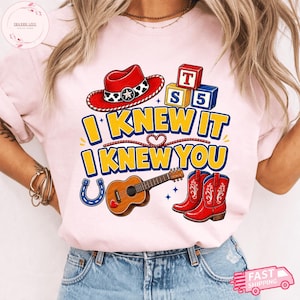 May include: A light pink t-shirt featuring a graphic print with a red cowboy hat, alphabet blocks labelled T, S, and 5, a brown acoustic guitar, and red cowboy boots. The text reads I KNEW IT I KNEW YOU in yellow letters with blue outlines.