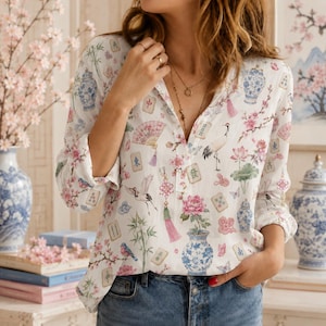 May include: A white long-sleeve button-down shirt featuring a colorful print of blue and white porcelain vases, pink cherry blossoms, cranes, lotus flowers, bamboo, and mahjong tiles. The shirt has a V-neckline and is worn with blue denim jeans.