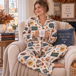 May include: A two-piece pajama set featuring a button-down long-sleeve shirt and matching pants with a cream background. The fabric is printed with autumn leaves, snowflakes, and icons including Luke's Diner, Doose's Market, and Stars Hollow signs.
