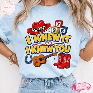 May include: A light blue t-shirt featuring a graphic print with a red cowboy hat, wooden alphabet blocks, a brown acoustic guitar, and red cowboy boots. The text reads I KNEW IT I KNEW YOU in yellow letters with blue outlines, accented by a horseshoe.