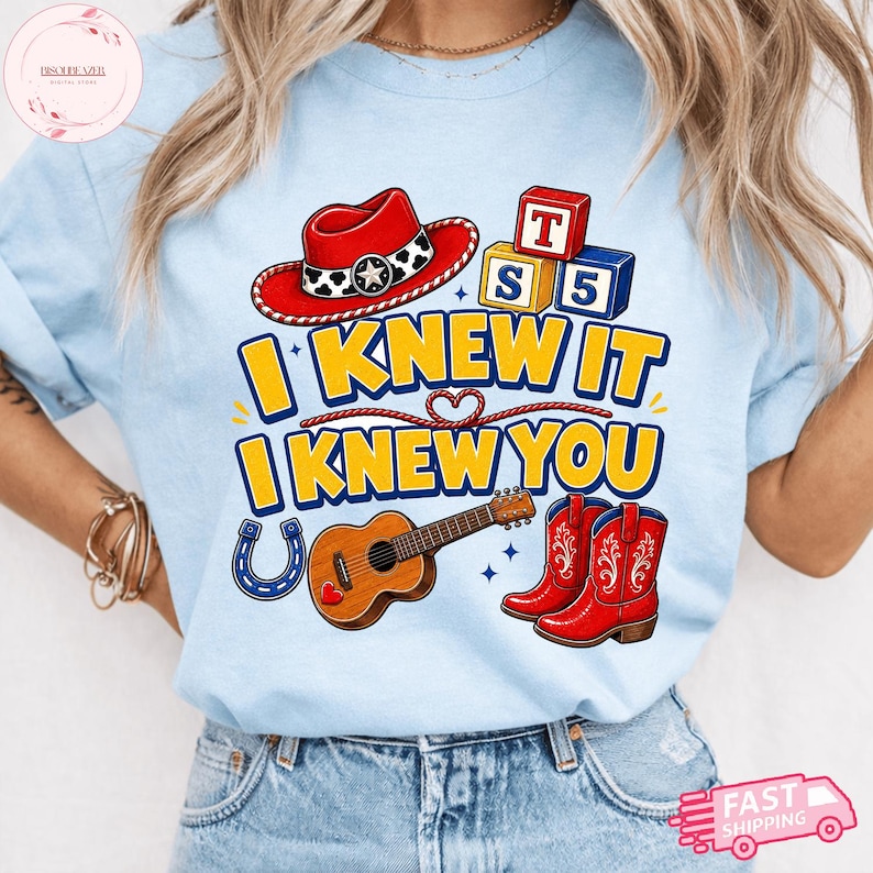 May include: A light blue t-shirt featuring a graphic print with a red cowboy hat, wooden alphabet blocks, a brown acoustic guitar, and red cowboy boots. The text reads I KNEW IT I KNEW YOU in yellow letters with blue outlines, accented by a horseshoe.