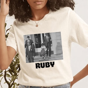 Könnte beinhalten: Cremefarbenes T-Shirt mit einem Schwarz-Weiß-Bild von Ruby Bridges. Das Bild ist zentriert und trägt das Wort "RUBY" in fetten, schwarzen Buchstaben darunter. Das T-Shirt besteht aus einem weichen Material.