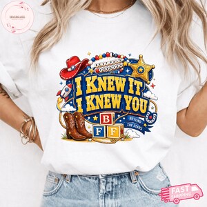 May include: A white t-shirt featuring a colorful graphic with the text I KNEW IT I KNEW YOU, BEST PALS FOREVER, and BEYOND THE STARS. The design includes a red cowboy hat, a gold sheriff star, a rocket, brown cowboy boots, and alphabet blocks B F F.