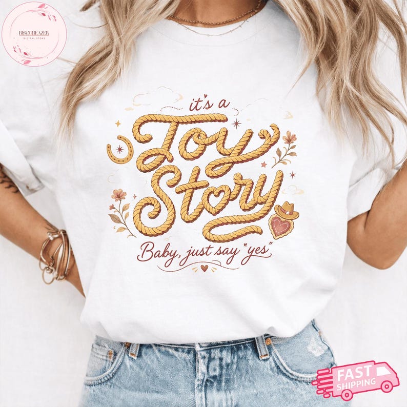 May include: A white t-shirt featuring a graphic design with the text "it's a Toy Story" written in a rope-like font, accompanied by the phrase "Baby, just say 'yes'" below, along with illustrations of a horseshoe, flowers, and a heart with a cowboy hat.
