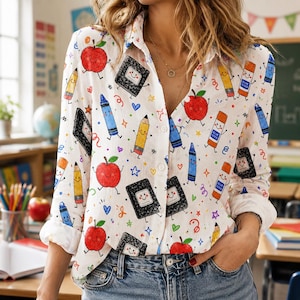 Pode incluir: Uma camisa branca de manga comprida com botões, apresentando um padrão colorido e repetitivo de material escolar desenhado, incluindo maçãs vermelhas, lápis amarelos, giz de cera azul, cadernos pretos e bastões de cola laranja com rostos.
