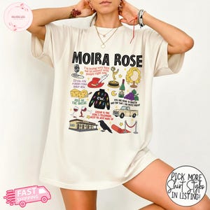 May include: Cream-colored t-shirt featuring the text "MOIRA ROSE" above a collection of illustrated icons. The illustrations include a microphone, a red hat, a cheese wedge, a black sweater, a wine bottle, and a vintage car. The shirt also has the text "FAST SHIPPING".