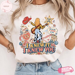 Może przedstawiać: Kremowy t-shirt z nadrukiem graficznym przedstawiającym osobę z długimi blond włosami grającą na gitarze, otoczoną czerwonym kowbojskim kapeluszem, gwiazdą, kwiatami i butami. Tekst brzmi "TAYLOR", "TS", "INFINITY AND BEYOND", "YOU'VE GOT A FRIEND IN ME", "I KNEW IT, I KNEW YOU".