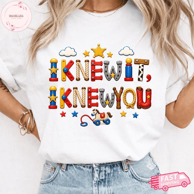 Puede incluir: Una camiseta blanca con un gr&aacute;fico colorido que incluye el texto I KNEW IT, I KNEW YOU. Las letras est&aacute;n dise&ntilde;adas para parecer varios juguetes y objetos de estilo western, incluyendo cuerdas, herraduras, estrellas y un caballito de juguete.