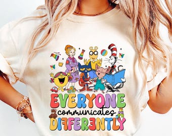 May include: Cream-colored t-shirt featuring a colorful graphic of various cartoon characters, including Mr. Men, Arthur, and The Cat in the Hat. The text "EVERYONE communicates DIFFERENTLY" is displayed in a rainbow font. A teddy bear and building blocks are also included in the design.
