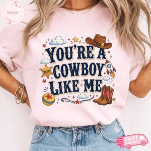 May include: A light pink t-shirt featuring the dark blue text "YOU'RE A COWBOY LIKE ME" surrounded by illustrations of a brown cowboy hat, a sheriff star badge, a rocket ship, a colourful ball, and cowboy boots, accented with small clouds and stars.