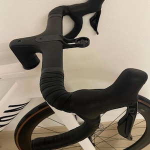 Può includere: Primo piano di un manubrio da bicicletta nero con leve freno integrate e nastro manubrio avvolto. La ruota ha un cerchio marrone e la scritta "DT SWISS". La bici è bianca.