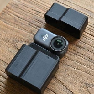 Puede incluir: Una pequeña cámara de acción gris oscuro con una lente visible, colocada entre dos estuches protectores negros. La cámara y los estuches están sobre una superficie de madera envejecida, lo que sugiere fotografía de aventura o al aire libre.