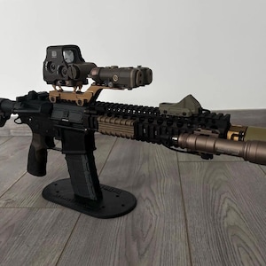 May include: A black and tan rifle with various attachments, including a scope, laser sight, and flashlight. The rifle is displayed on a black stand, highlighting its tactical design. The image focuses on the firearm's features.