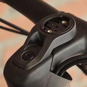 Puede incluir: Primer plano de una potencia de manillar de bicicleta negra con un soporte integrado para un ordenador de ciclismo. La potencia tiene un diseño elegante con la marca "Syncros" visible. El soporte tiene dos orificios para tornillos.